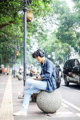 Smiling Young Asian Student Man Wearing Headphone Sitting on Sidewalk While Using Smartphone