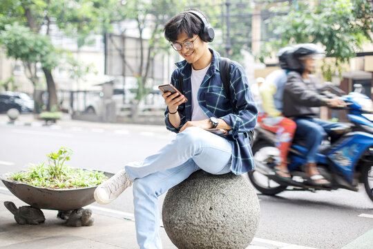 Smiling Young Asian Student Guy in Casual Style Sitting on Sidewalk While Using Smartphone