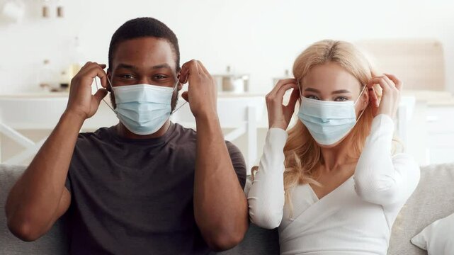A loving couple smiles while adjusting their masks at home. They embrace their multicultural relationship with warmth and affection while prioritizing health together.