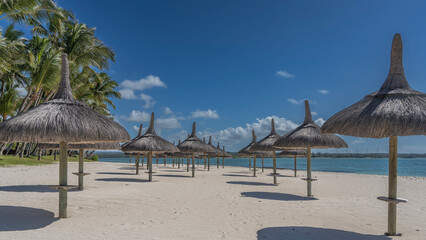 Rows of straw umbrellas on a tropical beach. Round shadows on white sand. Turquoise ocean. Palm trees against a background of blue sky and clouds. Mauritius. Resort.