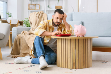 Stressed young man with calculator and piggy bank counting money at home