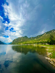 Stunning landscape featuring a calm lake reflecting the cloudy sky. Majestic mountains rise in the distance, framed by lush greenery, creating a tranquil and picturesque scene. Lake Idro, Crone, Italy