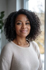 a happy, middle aged black woman with curly hair, wearing casual, standing by a window in a home office, looking into the camera and smiling