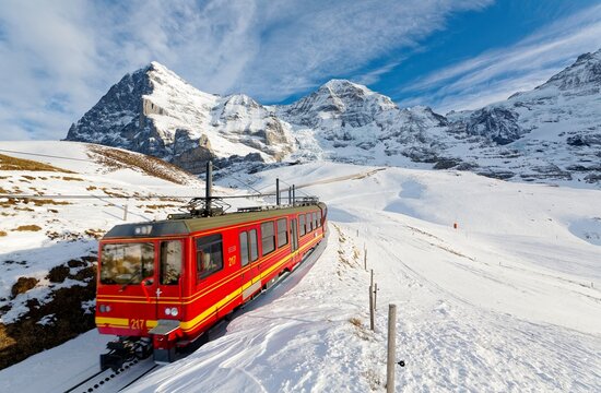 On a sunny winter day, tourists ride on a cogwheel train from Jungfraujoch (Top of Europe) to Kleine Scheidegg on the snowy hillside with Eiger and Monch in background, in Berner Oberland, Switzerland