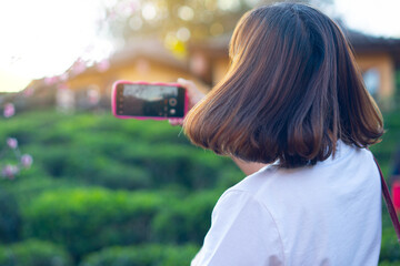 Women with shoulder length brown hair and is flowing. Wearing a white shirt. Takes a photo with a smartphone. The background is blurred greenery and a building.