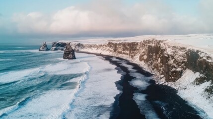 Stunning aerial winter landscape of black sand beach with ocean waves and snow-covered cliffs along the coast captured from a drone