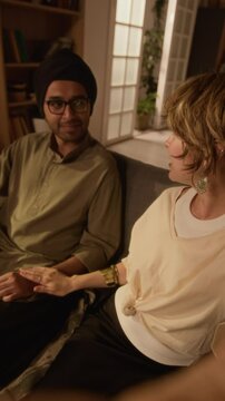 Vertical handheld UGC shot of young adult Caucasian blonde lady holding phone and waving to camera, sitting next to Indian husband in turban and glasses, talking and smiling at home
