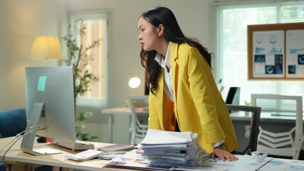 Professional asian businesswoman managing paperwork while working on desktop computer, focusing intently on corporate task in bright, modern office environment