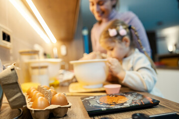 Close up of eggs and ingredients on kitchen counter with mother and daughter making cookies in blurry background.