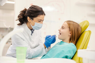 Brave girl sitting in char at dentist office with mouth opened while dentist doing teeth checkup.