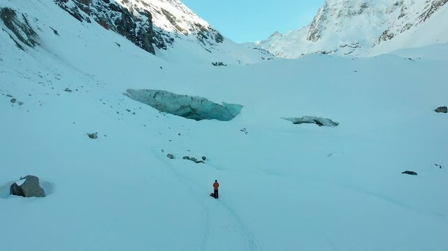 Aerial flyover of a winter hikers exploring a remote snow covered mountain valley of the Swiss Alps with a giant glacier cave in Valais, Switzerland during a cold winter day as the sun starts to set