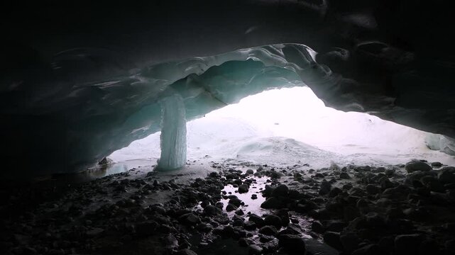 A winter adventurer with a camera climbs down a snow pactch to enter the darkness of a glacier cave in the Swiss Alps to inspect the ice of a giant frozen icicle and take photos