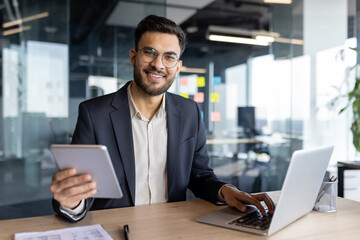 Portrait of a young successful businessman at the workplace, a man works with a laptop, sitting at a desk, holding a tablet computer in his hands. The employee is smiling and looking at the camera.