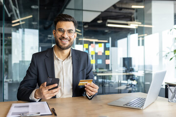 Portrait of young businessman inside office, man with bank credit card smiling and looking at camera. Office worker using laptop for online purchase and booking services.