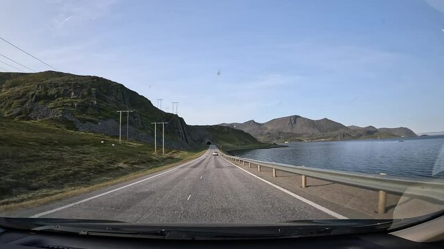 Driving through the tundra landscape of northern Norway on the E69 road. Rocky hills with grass surround the road. The sea is visible on the right side. The video includes a tunnel passage.