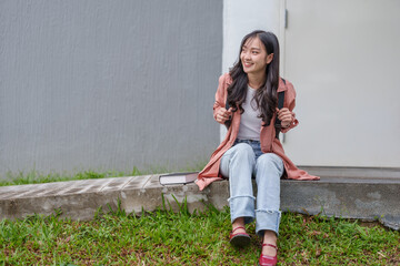 Happy asian female student wearing casual clothing, sitting on a step in front of a university...