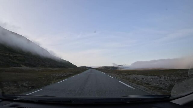 Driving through the tundra in northern Norway on E69 road. Grasslands and a rocky hill surround the area. Low clouds create a serene and atmospheric view of the Arctic landscape.