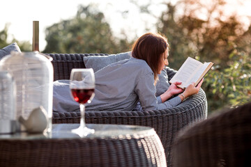A woman is relaxing on the veranda of a country house and reading a book over a glass of red alcoholic drink or wine. The concept of relaxation and luxury wine.