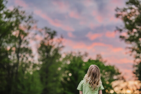 Child watching a vibrant pink and orange sunset in a forest clearing.
