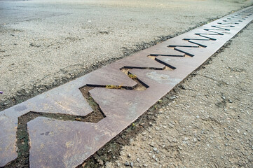 A close up view of a bridge expansion joint with a zigzag pattern, designed to accommodate structural movements and prevent damage. The metal plate is embedded in the asphalt road surface