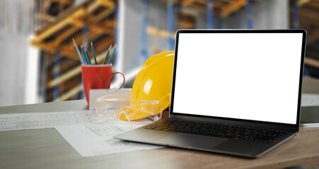 A laptop is open on a desk with a yellow hard hat and a red cup of pens. working table architect...