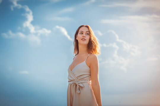 young brunette woman standing, wearing a cream white dress with lightly clouded sky