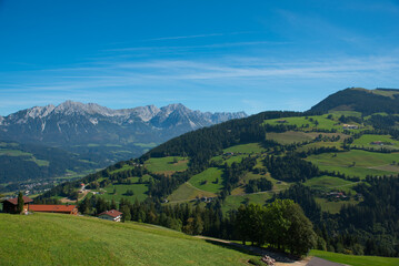 A breathtaking Alpine landscape with lush green meadows, forested hills, and distant rocky peaks. A small artificial pond and building in the foreground.