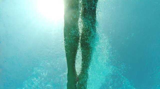 View from bottom of pool to young man diving into crystal clear water. Guy refreshing in basin during resting on resort. Male tourist enjoying summer vacation. Underwater footage. Slow mo Close up