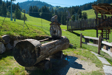 A whimsical wooden sculpture fountain in an Alpine setting. A carved face spouts water into a hollowed log. Surrounding it are green meadows, forested hills, and a wooden playground structure.