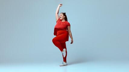 Smiling chubby woman wearing red t-shirt, red leggings, white sneakers, standing mid-dance with hands high against beige studio background. Concept of beauty, fitness, movement, dance and music.
