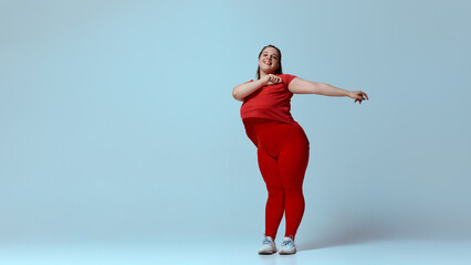 Dynamic portrait of smiling fat woman dressed red activewear, dancing energetic jazz-funk in motion against blue studio background. Concept of beauty, fitness, movement, dance and music.