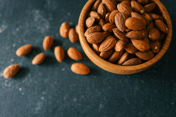 Rustic wooden bowl filled with raw almonds placed on a dark textured surface. Scattered almonds add...