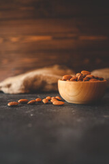 Rustic wooden bowl filled with raw almonds placed on a dark textured surface. Scattered almonds add a natural touch, complemented by a burlap fabric in the background