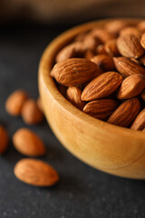 Rustic wooden bowl filled with raw almonds placed on a dark textured surface. Scattered almonds add a natural touch, complemented by a burlap fabric in the background