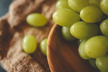 Close-up of fresh green grapes in a wooden bowl on a textured burlap fabric. Rustic and natural food concept with a soft, warm aesthetic