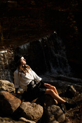 a woman sitting on the rocks next to a small waterfall. She is wearing a white shirt and a black skirt, as well as black boots. She has a black hat on her head.