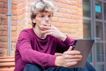 Young man in a burgundy shirt sitting against a brick wall, holding a tablet and looking thoughtful, outdoors in an urban setting © Raquel