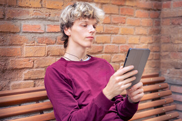 Young man in a burgundy shirt sitting against a brick wall, holding a tablet and looking thoughtful, outdoors in an urban setting © Raquel