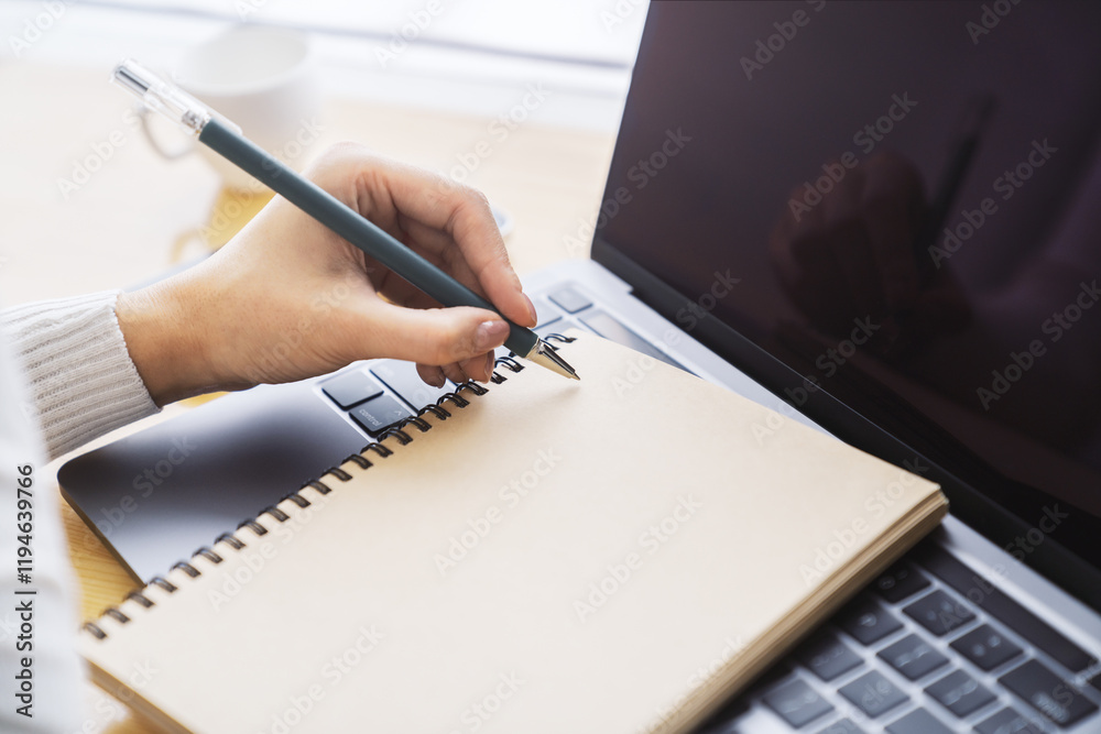 Poster close-up depiction of a woman's hand taking notes in a notepad on a sleek laptop, with a blurred bac