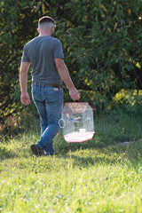 A man walks across a grassy area, carrying a small birdcage. The birdcage contains a small bird....