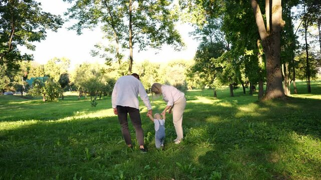Young parents walk with toddler on green grass in park in summer. Family helps child to take first steps and develop walking skills. Concept of skills development, support, childhood, family. 