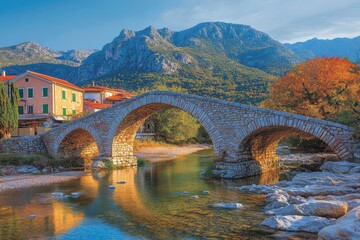 Naklejka premium A vibrant scenic view featuring a stone bridge arching over a calm river, with colorful houses on one side and majestic mountains as a striking backdrop, under a clear blue sky.