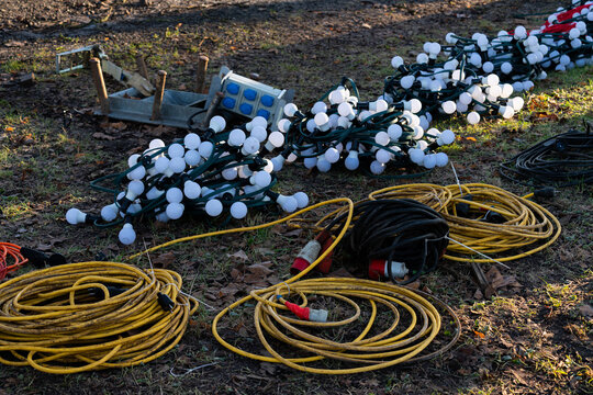 Numerous strings of decorative lights and extension cords lay on the ground in a park setting, indicating preparations for an outdoor event during daylight hours.