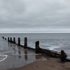 Fototapeta premium Weathered wooden groins extend into the sea beneath a cloudy sky, against a tranquil coastal scene.