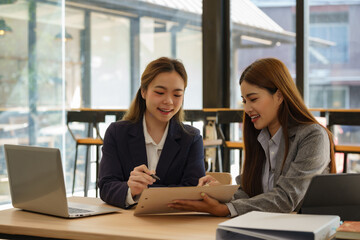Two Asian businesswomen sit at desks discuss project details, share opinions, and work on collaborative tasks.