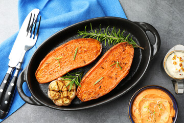 Halves of tasty cooked sweet potato with rosemary and garlic in baking dish served on grey textured table, flat lay