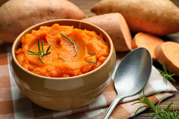Tasty mashed sweet potato with rosemary in bowl, fresh vegetables and spoon on table, closeup