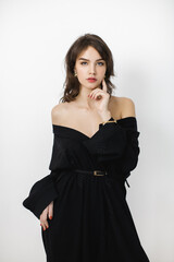 Fototapeta premium A beautiful brunette woman wearing a black off-shoulder dress and gold accessories poses confidently against a minimalist white background. Ideal for fashion, beauty, and elegance concepts. 