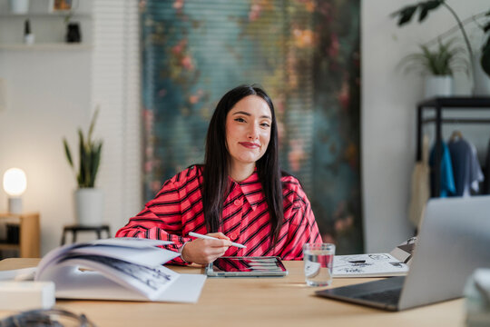 Creative individual wearing a striped shirt is focused on sketching on a tablet at a desk surrounded by drawing materials and a glass of water, showcasing a modern artistic workspace.