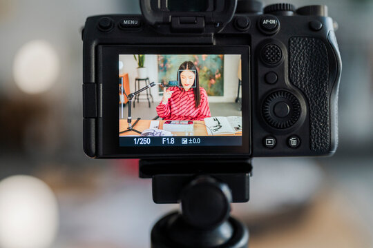 camera screen shows a digital artist in a striped pink shirt at a desk with sketchbooks and a tablet, framed by a professional recording setup in a modern studio.
